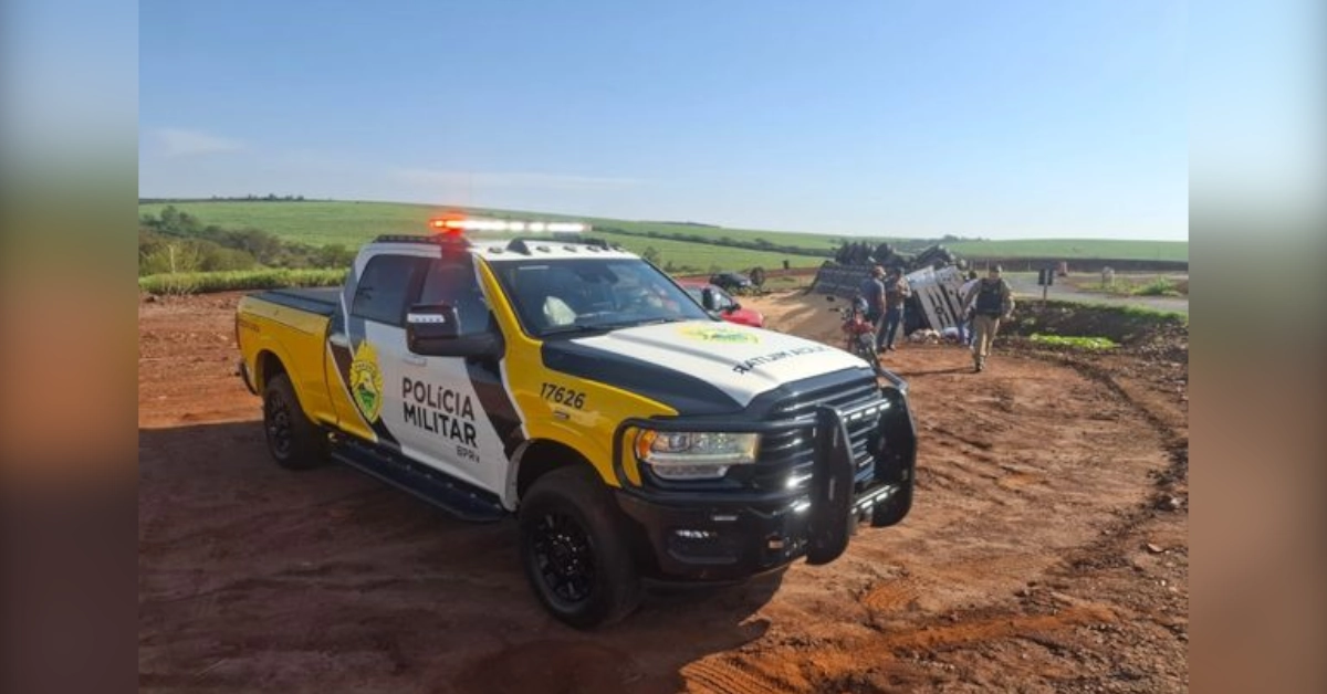 Caminhão tomba na PR-317 em Iguaraçu e deixa passageiro ferido. Imagem mostra viatura da Polícia Militar sinalizando o trecho da rodovia ao lado do veículo tombado.